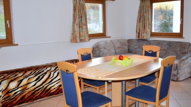 Uma área de refeições com uma mesa redonda, quatro cadeiras, um sofá e duas janelas com cortinas.