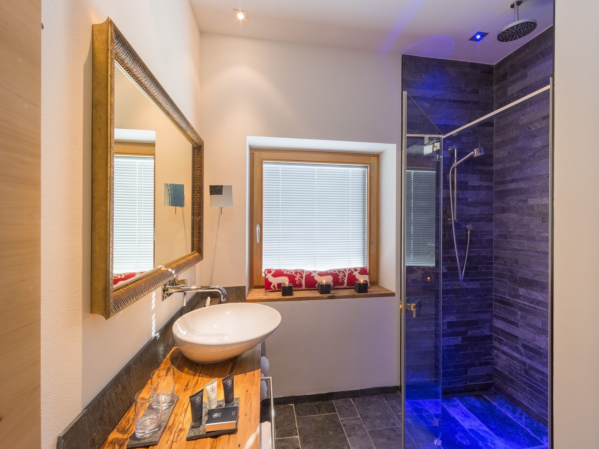 A bathroom with a large mirror, a sink, a window with blinds, and a shower area with a dark tiled wall.