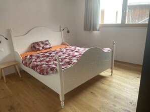 Une chambre avec un lit simple, une tête de lit blanche, un plancher en bois, une petite table de chevet et une fenêtre donnant sur l’extérieur.