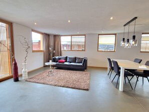 A modern living room with a sofa, a dining table with chairs, a decorative vase, and large windows.