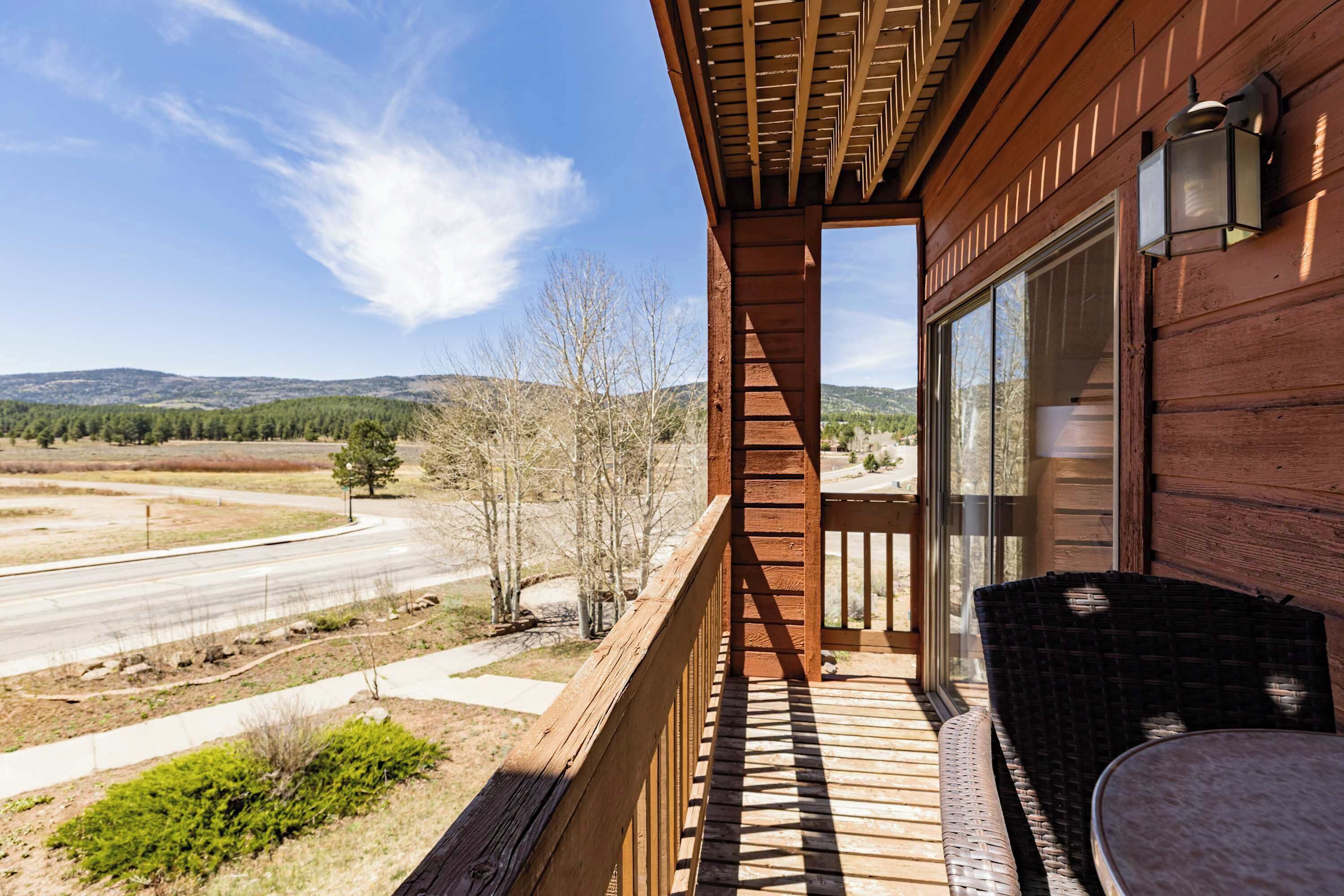Una terraza de madera con vista a una carretera y colinas, una silla de mimbre y una mesa.