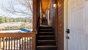 A wooden staircase leading to a white door, with a view of a parking area and trees in the background. - Evergreen Escape by Velo (Angel Fire)