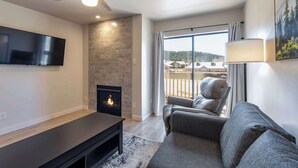 A modern living room with a fireplace, a flat-screen TV, a ceiling fan, and a large window with a view of houses. - Evergreen Escape by Velo (Angel Fire)