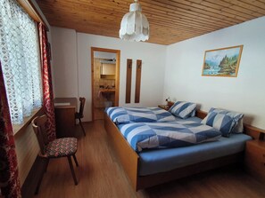 Ein Schlafzimmer mit Bett, Schreibtisch, Stuhl, einem Fenster mit Vorhängen, Holzboden und einem Bild an der Wand.