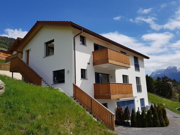 A two-story building with a balcony on the second floor, a staircase leading to the entrance, and a view of mountains in the background. - Chasa Scandella, (Sent) by Interhome (Sent)