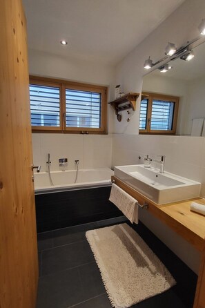A modern bathroom with a bathtub, a double sink, and a large mirror. - Chasa Scandella, (Sent) by Interhome (Sent)