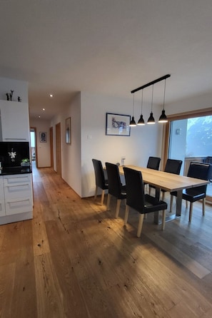 A modern dining area with a wooden table and black chairs, a kitchen with white cabinets, and a framed picture on the wall. - Chasa Scandella, (Sent) by Interhome (Sent)