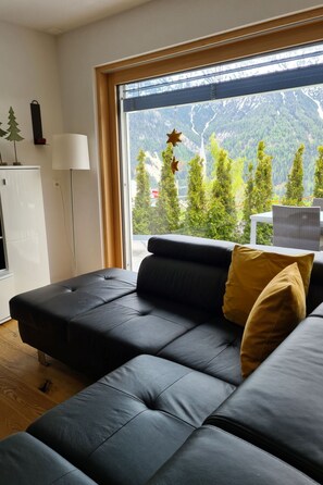 Ett modernt vardagsrum med en svart lädersoffa, en golvlampa och utsikt över ett bergigt landskap genom ett stort fönster.