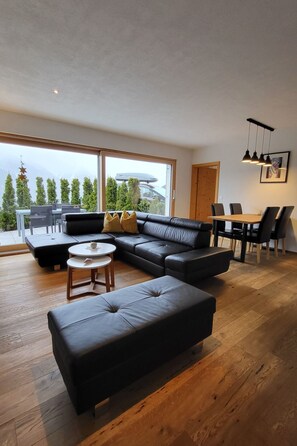 A modern living room with a black leather sofa, a round coffee table, and a dining area with a wooden table and chairs. - Chasa Scandella, (Sent) by Interhome (Sent)