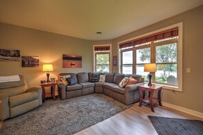 Ein Wohnzimmer mit einem Ecksofa, zwei Beistelltischen mit Lampen, einem Teppich und großen Fenstern mit Jalousien.