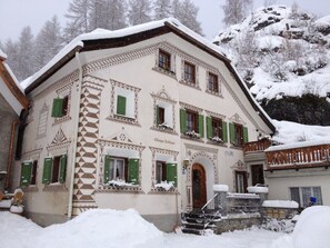 A traditional building with a snow-covered exterior, multiple windows with shutters, and a staircase leading to the entrance.