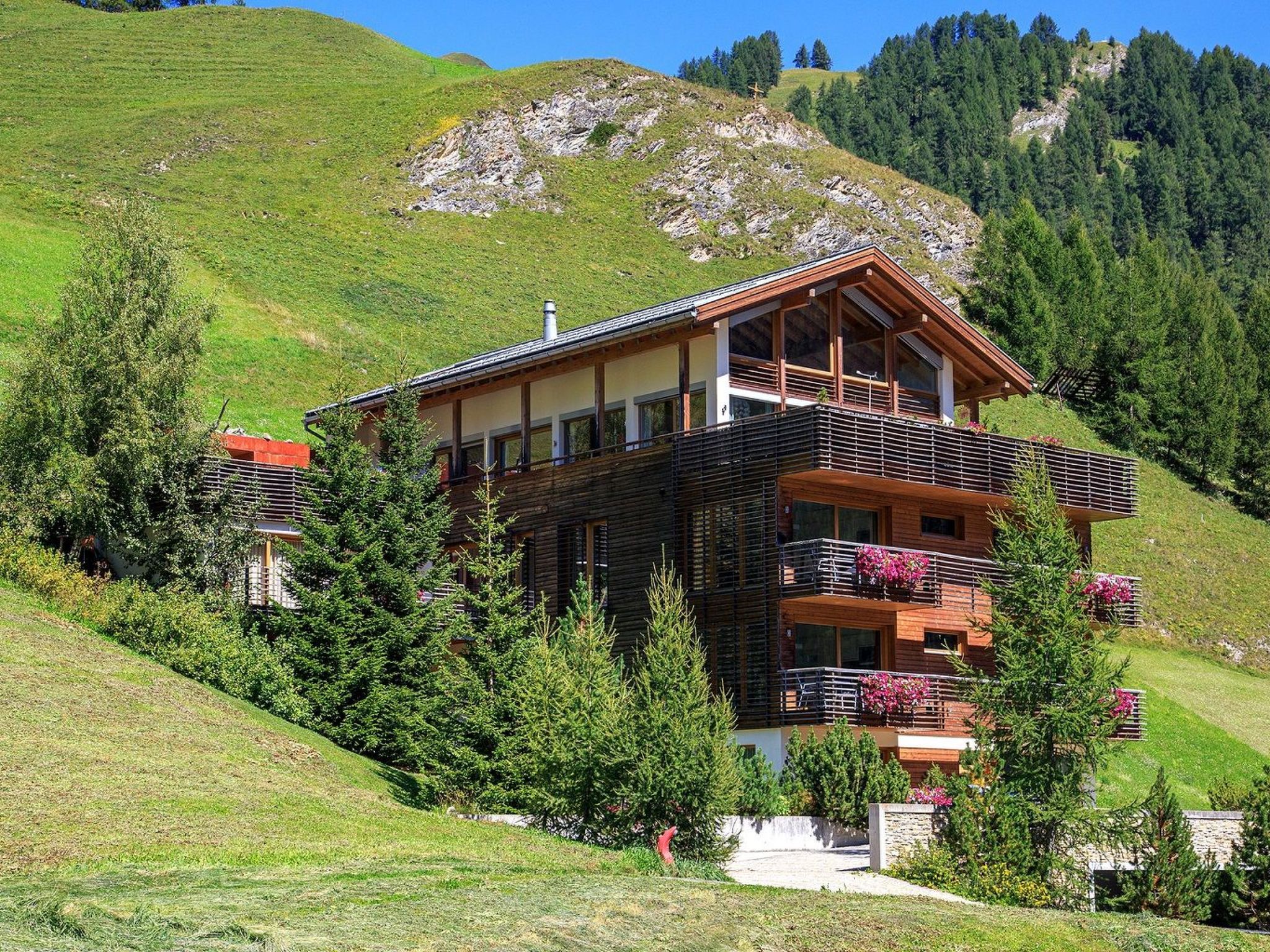 Un chalet à plusieurs étages doté d’un balcon, entouré de verdure et adossé à un paysage montagneux.