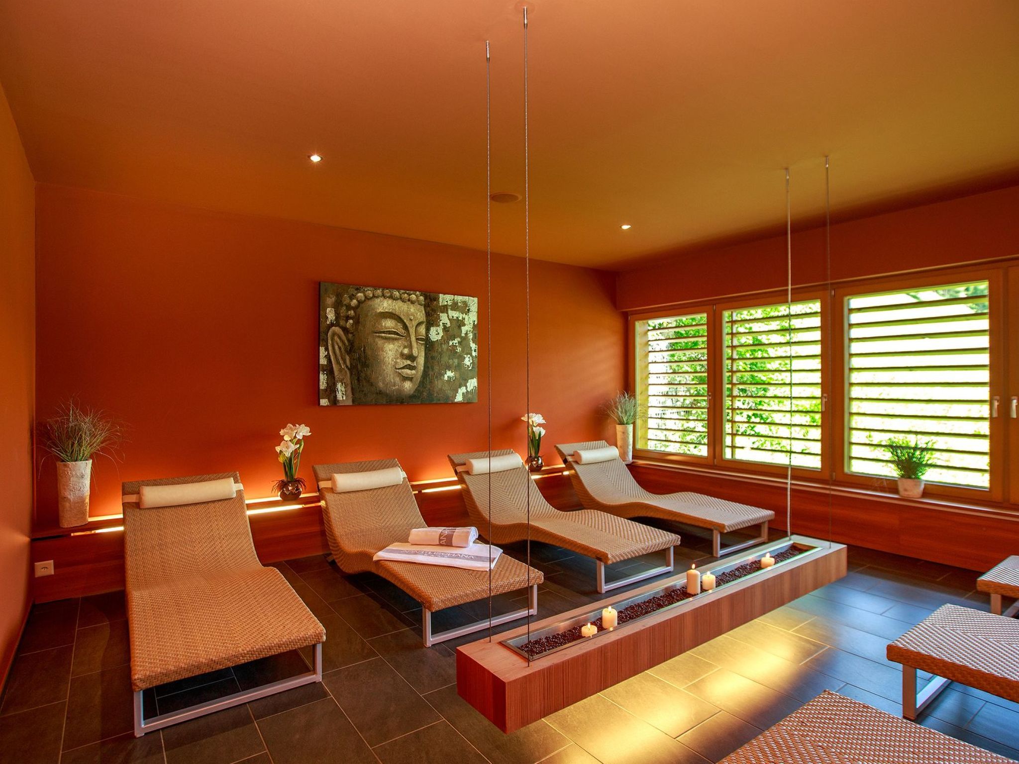 Ein moderner Wellnessraum mit Liegesesseln, einem großen Buddha-Gemälde und Blick auf Grün durch große Fenster.