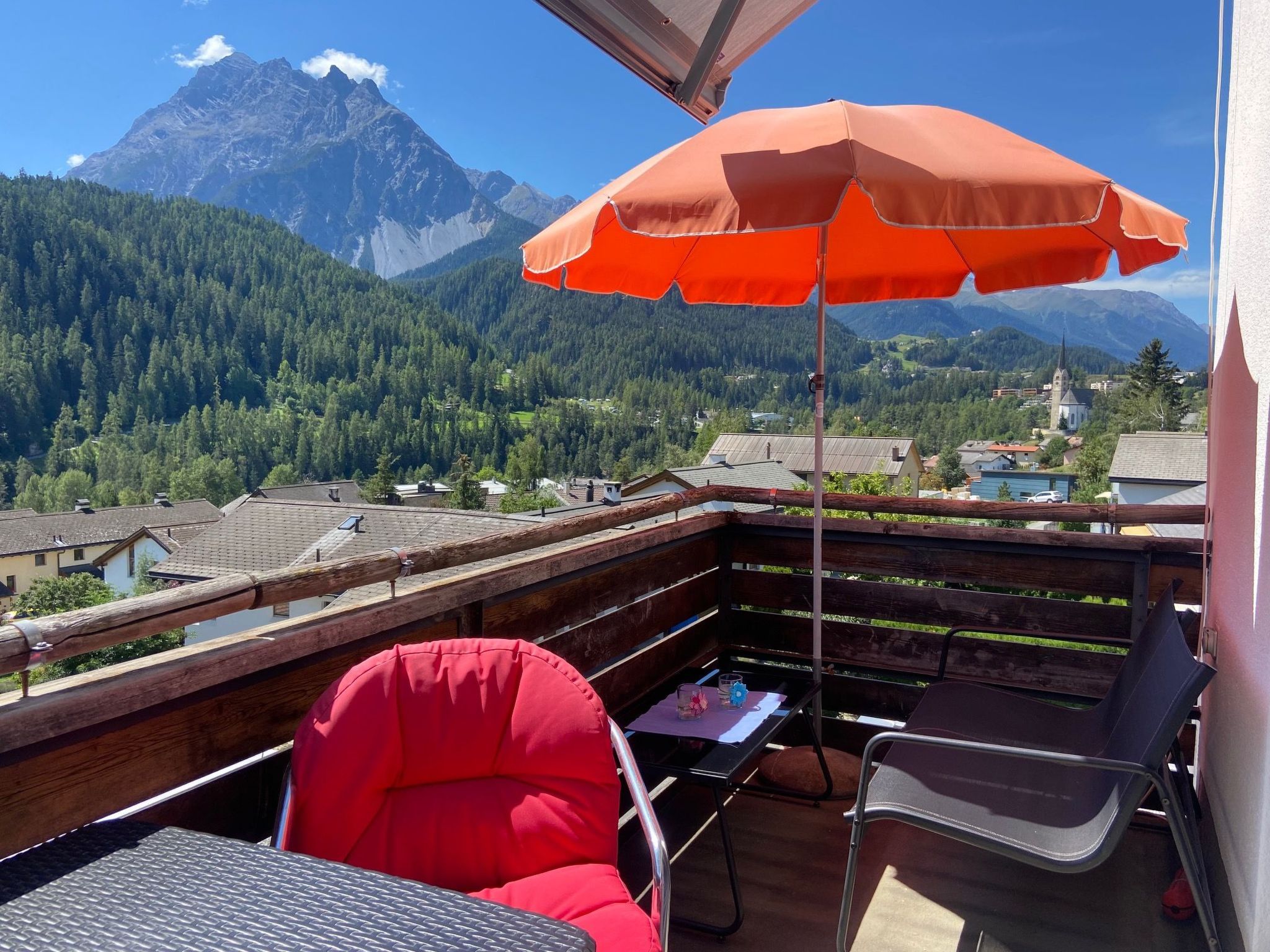 A balcony with a red umbrella, two chairs, and a table.