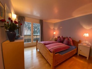 A bedroom with a wooden bed, two bedside lamps, a nightstand, a window with curtains, and a vase with flowers on a dresser. - Sotchà 8c, (Scuol) by Interhome (Scuol)