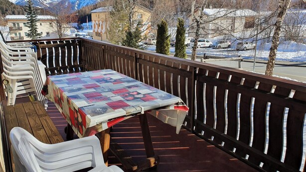 Een balkon met een tafel en stoelen, uitzicht op een besneeuwde berg en een parkeerplaats met auto's.