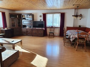 Een ruime woonkamer met een houten plafond, een televisie, een eethoek met een tafel en stoelen, en een boekenkast.