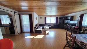 Een ruime woonkamer met een houten plafond, een eettafel met stoelen, een bank, een televisie en een keuken met kasten en een raam.