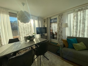 A hotel room with a dining area, a flat-screen TV, a sofa with cushions, and a large window with curtains. - Chasa Uleiva, (Samnaun-Compatsch) by Interhome (Samnaun)