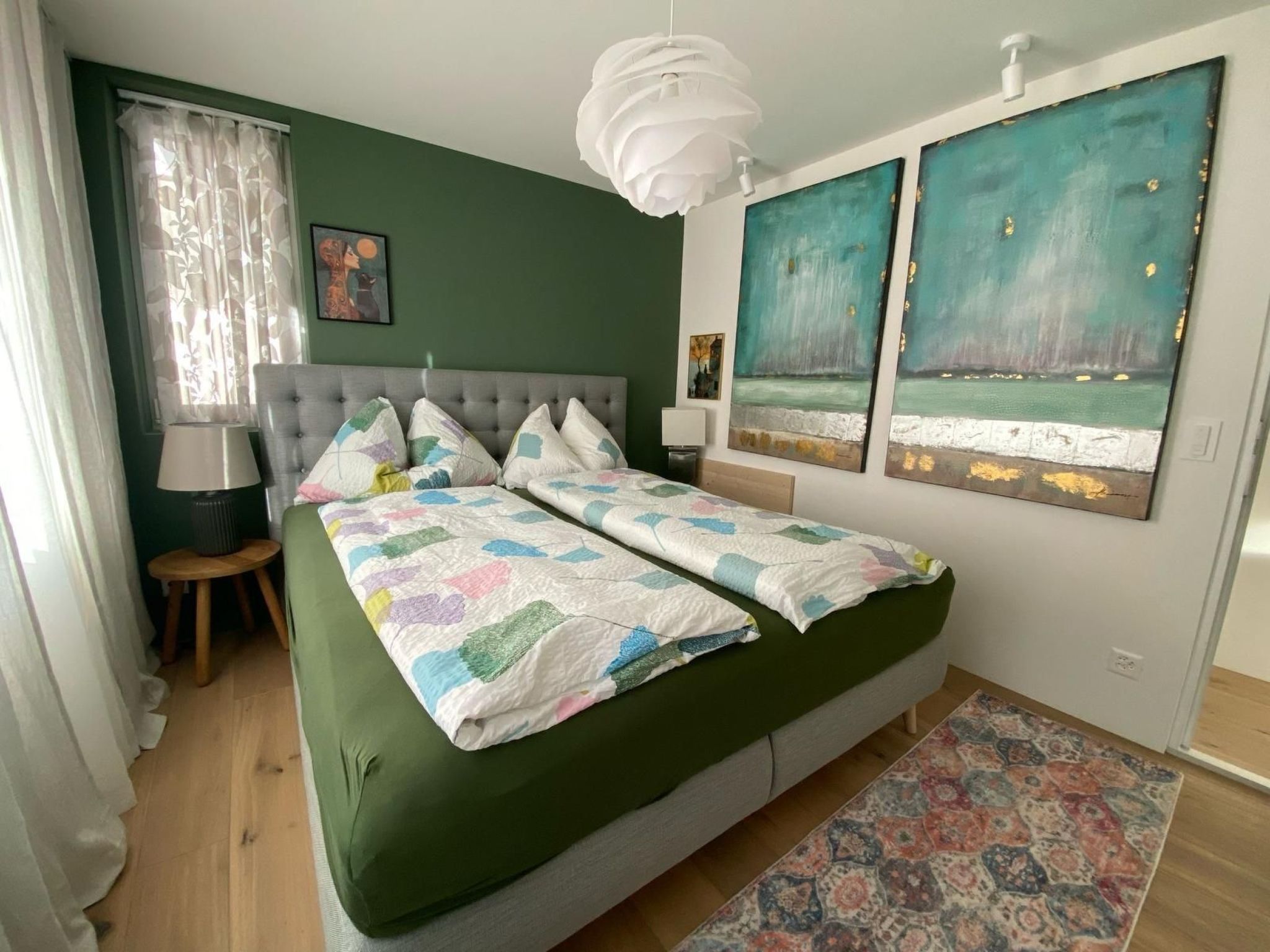 Ein Schlafzimmer mit einem großen Bett, einem Nachttisch, einer Lampe, einem Fenster mit Vorhängen und zwei Bildern an der Wand.