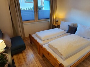 A double bed with white linens, a bedside table with a lamp, and a view of a blue building through a window with blinds. - Ferienwohung Chasa Max, (Samnaun-Plan) by Interhome (Samnaun)