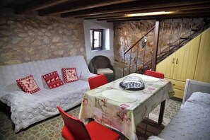 Una habitación acogedora con pared de piedra, una escalera de madera, un sofá blanco con cojines rojos, una mesa de comedor con mantel floral y sillas rojas.