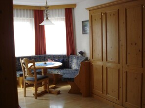 Ein Zimmer mit einem hölzernen Kleiderschrank, einem runden Tisch mit Stühlen, einem blauen, gemusterten Sofa und einem Fenster mit roten Vorhängen.