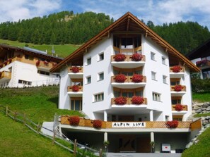 A multi-story hotel with balconies adorned with red flowers, set against a backdrop of green hills. - Appartements Alpin Live, (Samnaun-Laret) by Interhome (Samnaun)