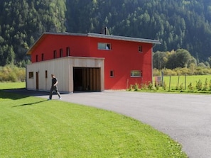 A red building with a garage door open, a person walking in front, and a green field with trees in the background.