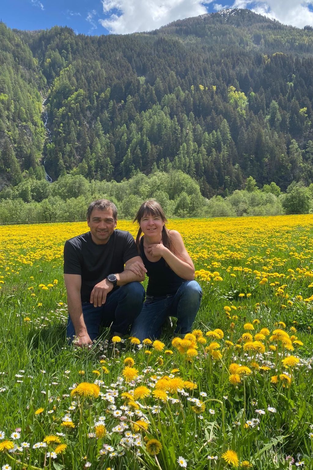 To personer sidder i en mark med gule blomster, med et skovklædt bjerg i baggrunden.