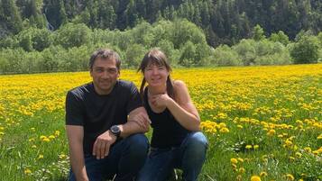 To personer sidder i en mark med gule blomster, med et skovklædt bjerg i baggrunden.
