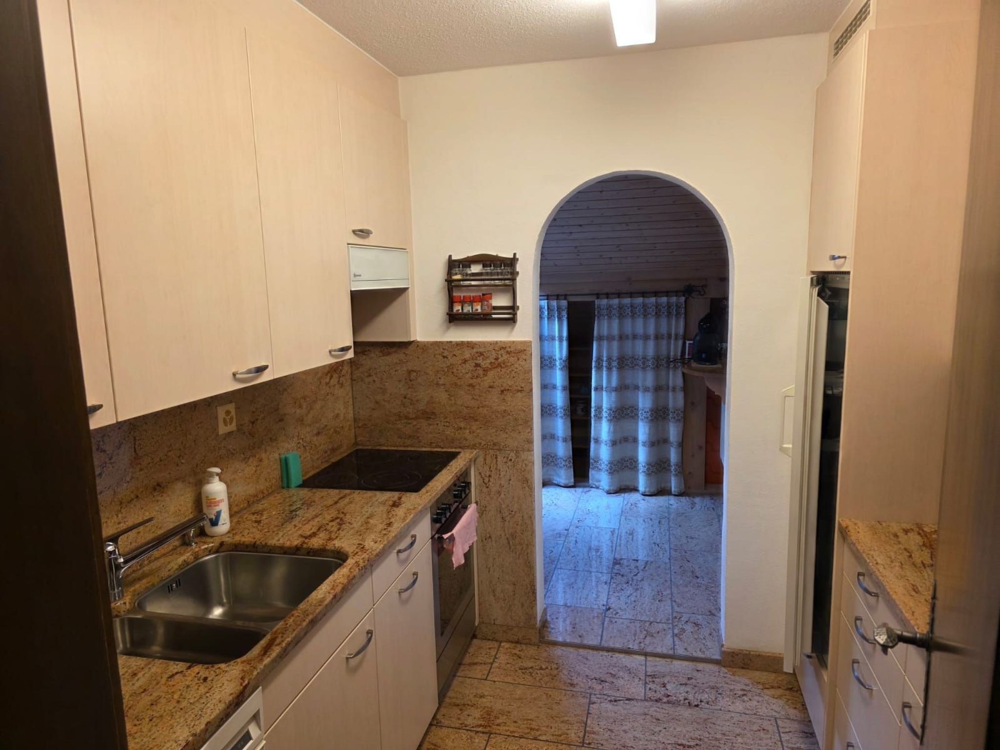 A kitchen with granite countertops, a sink, and cabinets. There is a small shelf with kitchen items and a window with curtains.