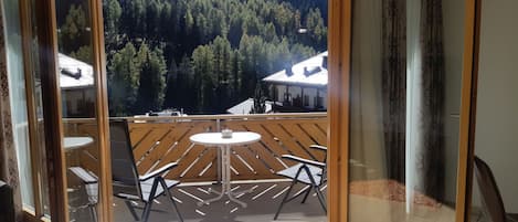 A balcony with a table and chairs, a view of a forested mountain, and a building with a sloped roof.