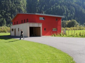 Um edifício vermelho com uma porta de garagem aberta, uma pessoa caminhando na frente e um campo verde com árvores ao fundo.