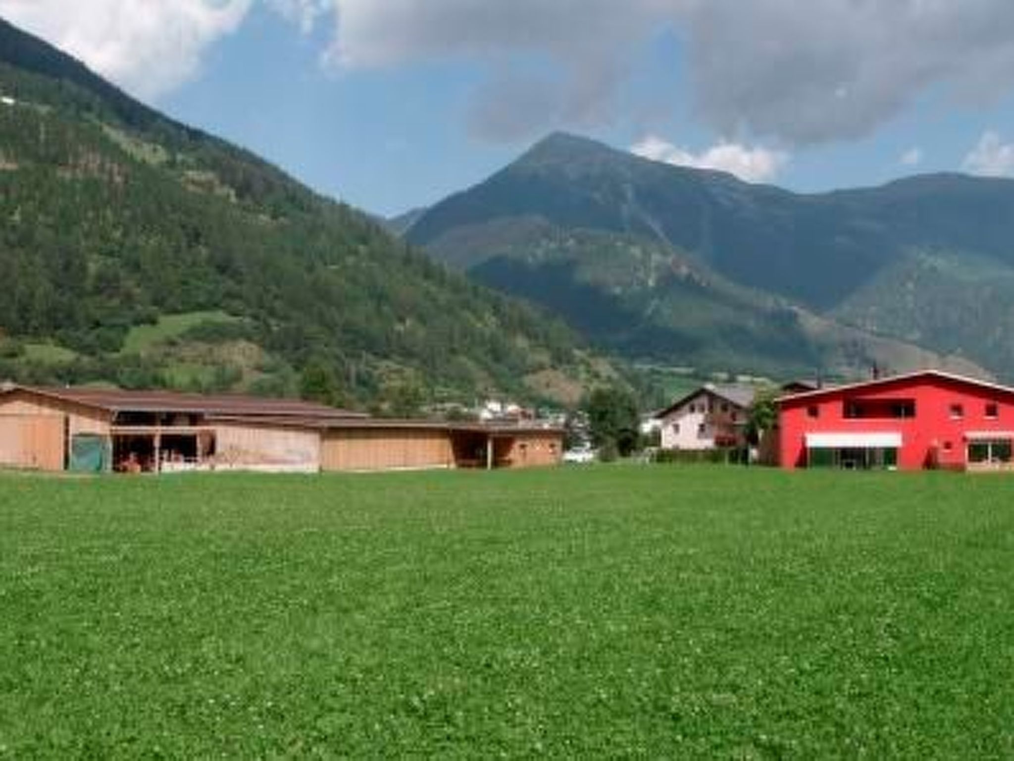 Ländliche Landschaft mit einem grünen Feld, zwei Gebäuden und Bergen im Hintergrund.