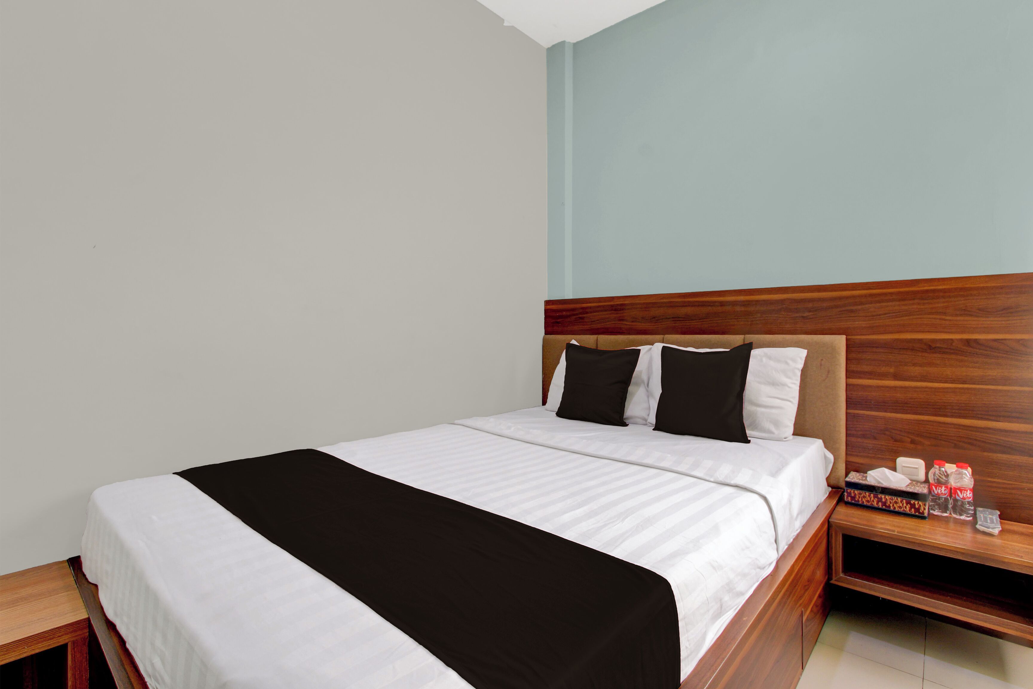 A neatly made bed with a wooden headboard, a bedside table with a bottle and a box, and a plain wall.
