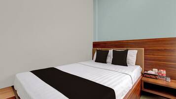 A neatly made bed with a wooden headboard, a bedside table with a bottle and a box, and a plain wall.