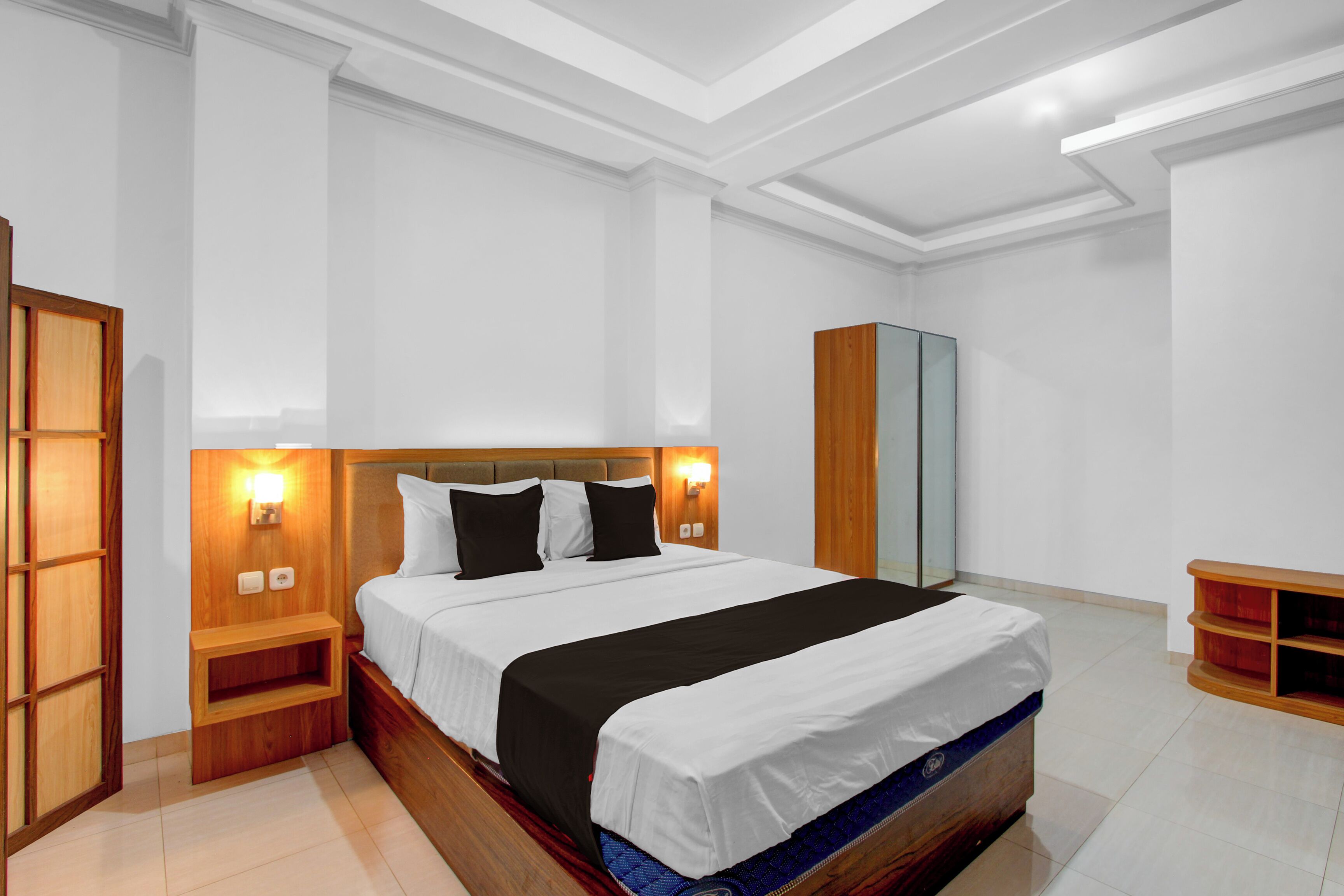 A modern hotel room with a large bed, wooden headboard, bedside tables, a wardrobe, and a built-in shelf.