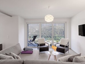Ein modernes Wohnzimmer mit einer Couch, zwei Sesseln, einem kleinen Tisch mit Schüssel und Gläsern sowie einer Pendelleuchte. Große Fenster bieten Blick auf ein bewaldetes Gebiet.