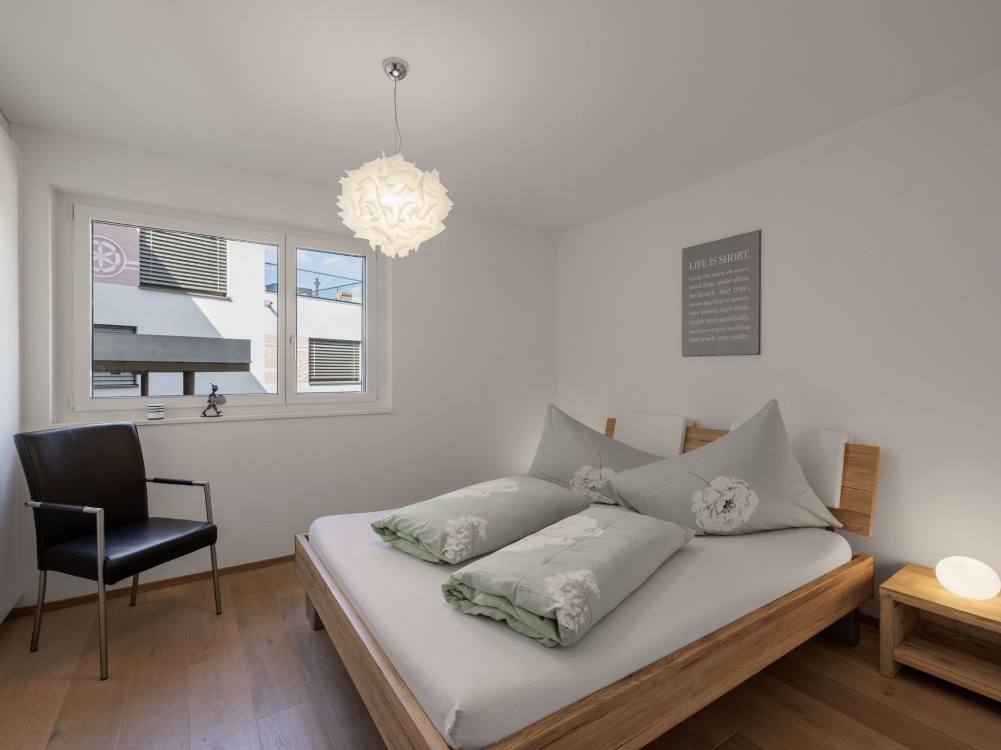 Ein Schlafzimmer mit Bett, Sessel, Nachttisch, Wanduhr, Fenster mit Jalousien und Deckenleuchte.