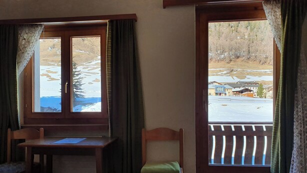 Ein Zimmer mit einem Bett, einem Schreibtisch und einem Stuhl. Zwei Fenster mit Vorhängen, eines davon bietet Blick auf eine verschneite Landschaft.