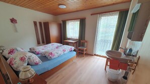 Ein Schlafzimmer mit Holzboden, einem Bett mit floralem Bettzeug, einem kleinen Tisch, einem Stuhl, einem Fenster mit Spitzenvorhängen und einem Badezimmer mit Waschbecken und Spiegel.