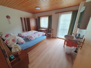 Ein Schlafzimmer mit Holzboden, einem Bett mit floralem Bettzeug, einem kleinen Tisch, einem Stuhl, einem Fenster mit Spitzenvorhängen und einem Badezimmer mit Waschbecken und Spiegel.