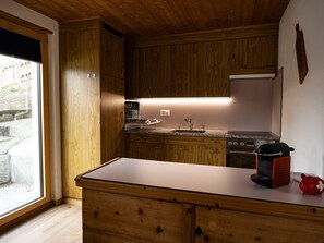 Cozinha com armários de madeira, geladeira de aço inoxidável, forno embutido e janela com vista para um edifício.
