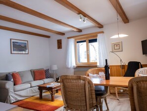 Uma sala de estar com sofá, mesa de jantar, cadeiras de vime, piso de madeira e uma janela com cortinas.