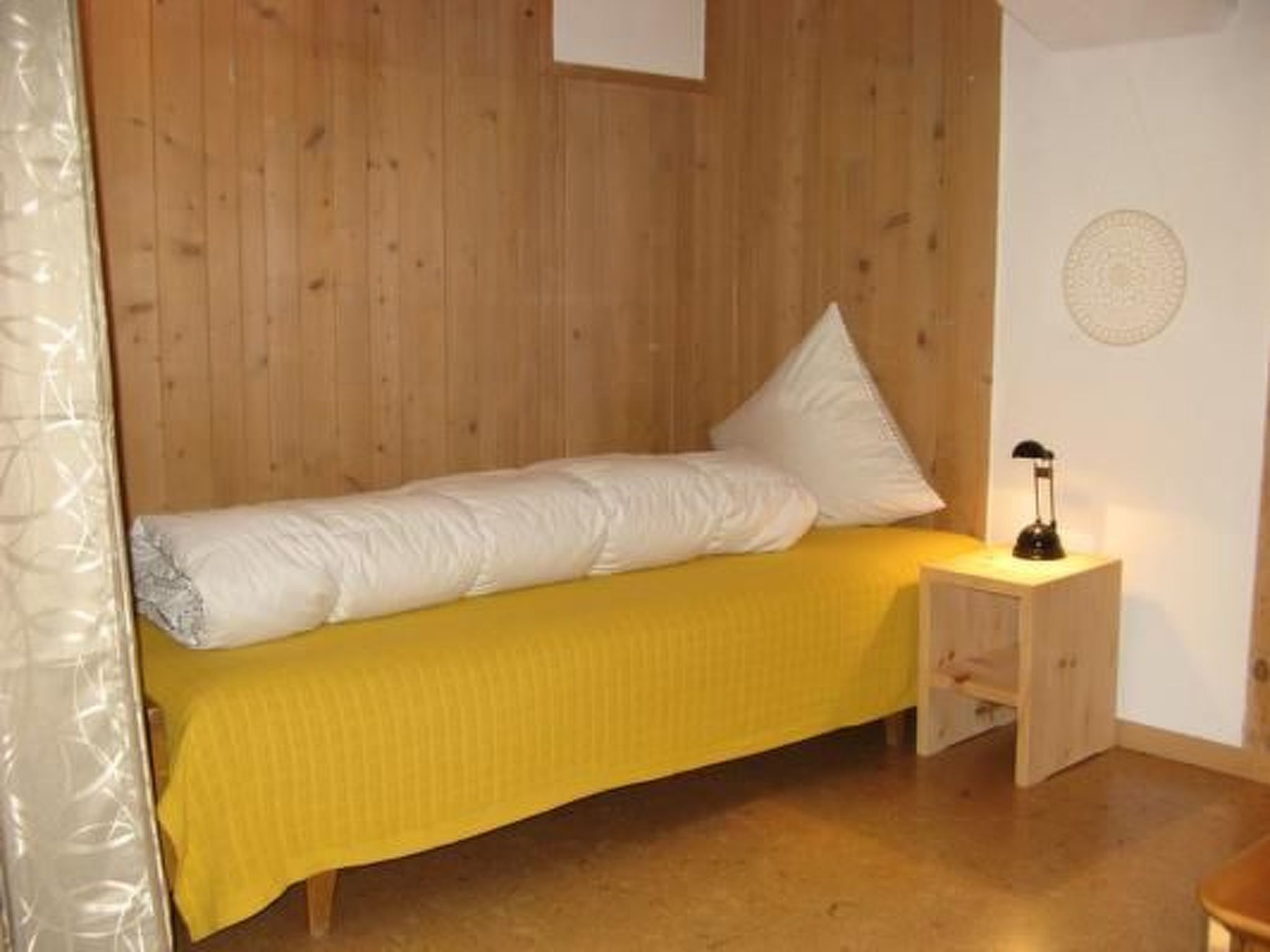 En seng med gul sengetæppe og hvide puder i et rum med trævægge og et lille natbord med en lampe.
