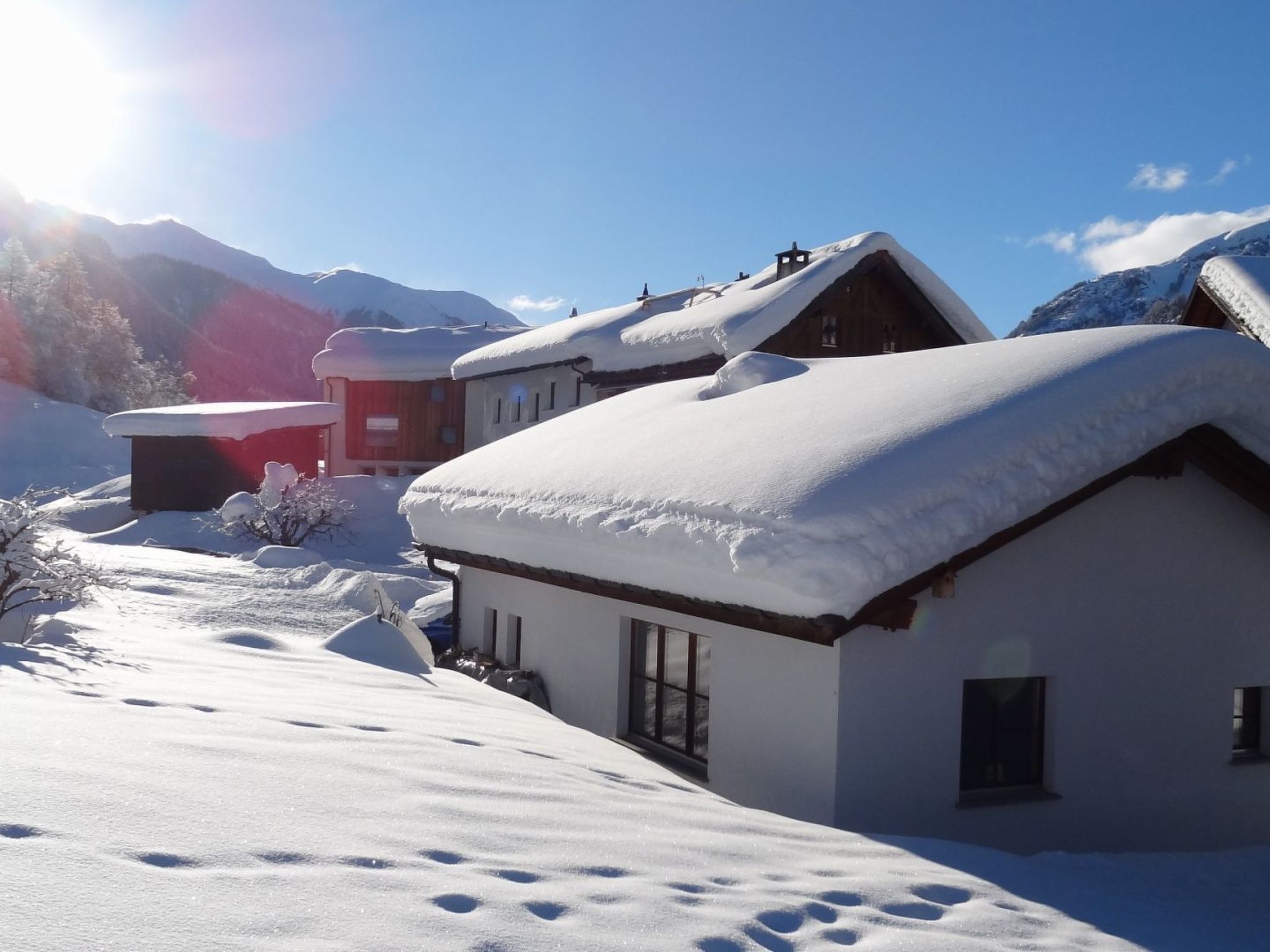 A snow-covered mountain village with traditional wooden houses, a clear sky, and a bright sun.