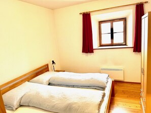 A small, single-bed room with a wooden headboard, a window with red curtains, and a bedside lamp.
