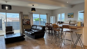 Un salón espacioso con sofá de cuero negro, mesa de comedor y sillas, una cocina con barra y amplios ventanales con vistas al exterior.