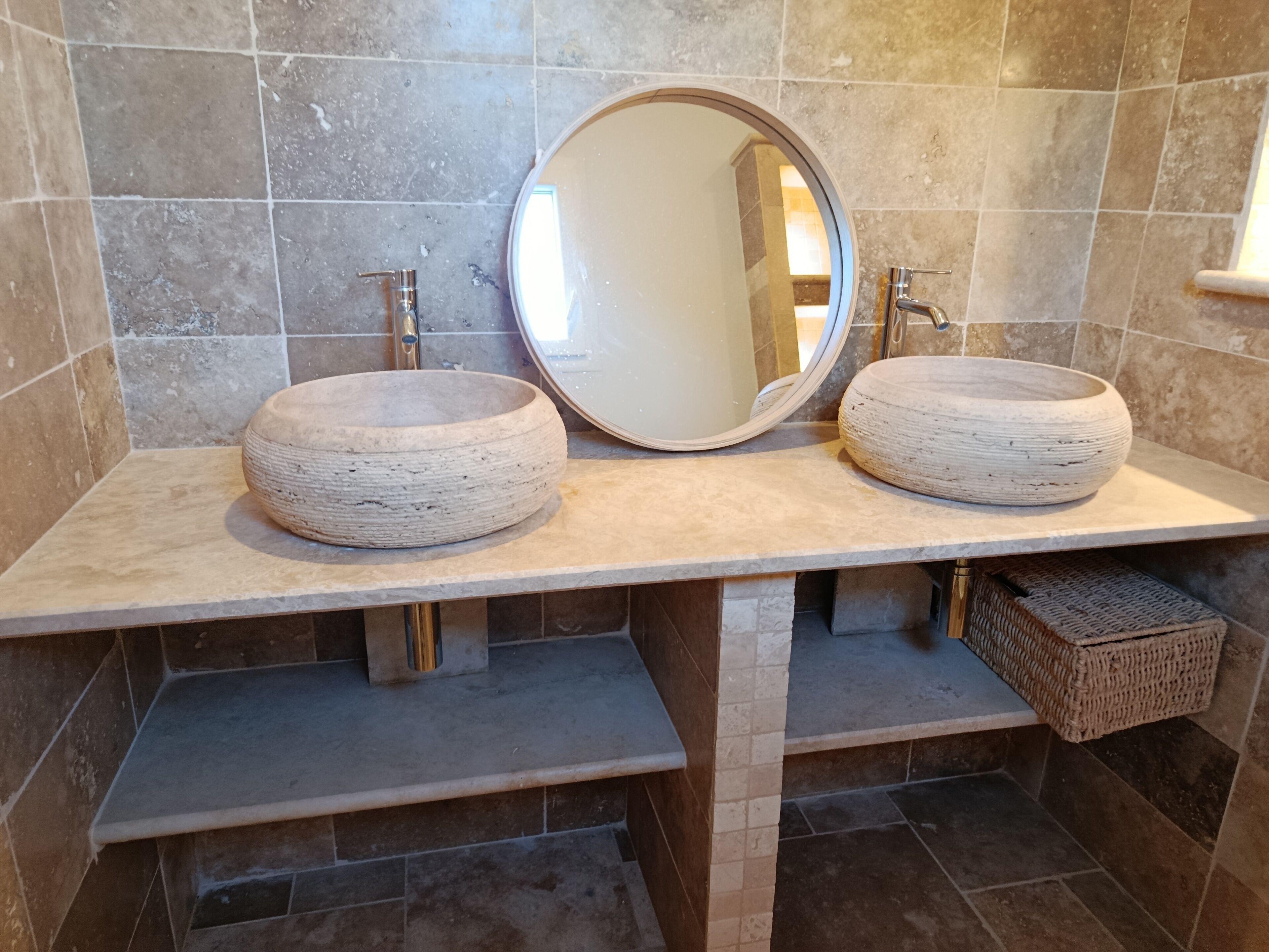 A bathroom with two stone sinks, a round mirror, and a wicker basket.